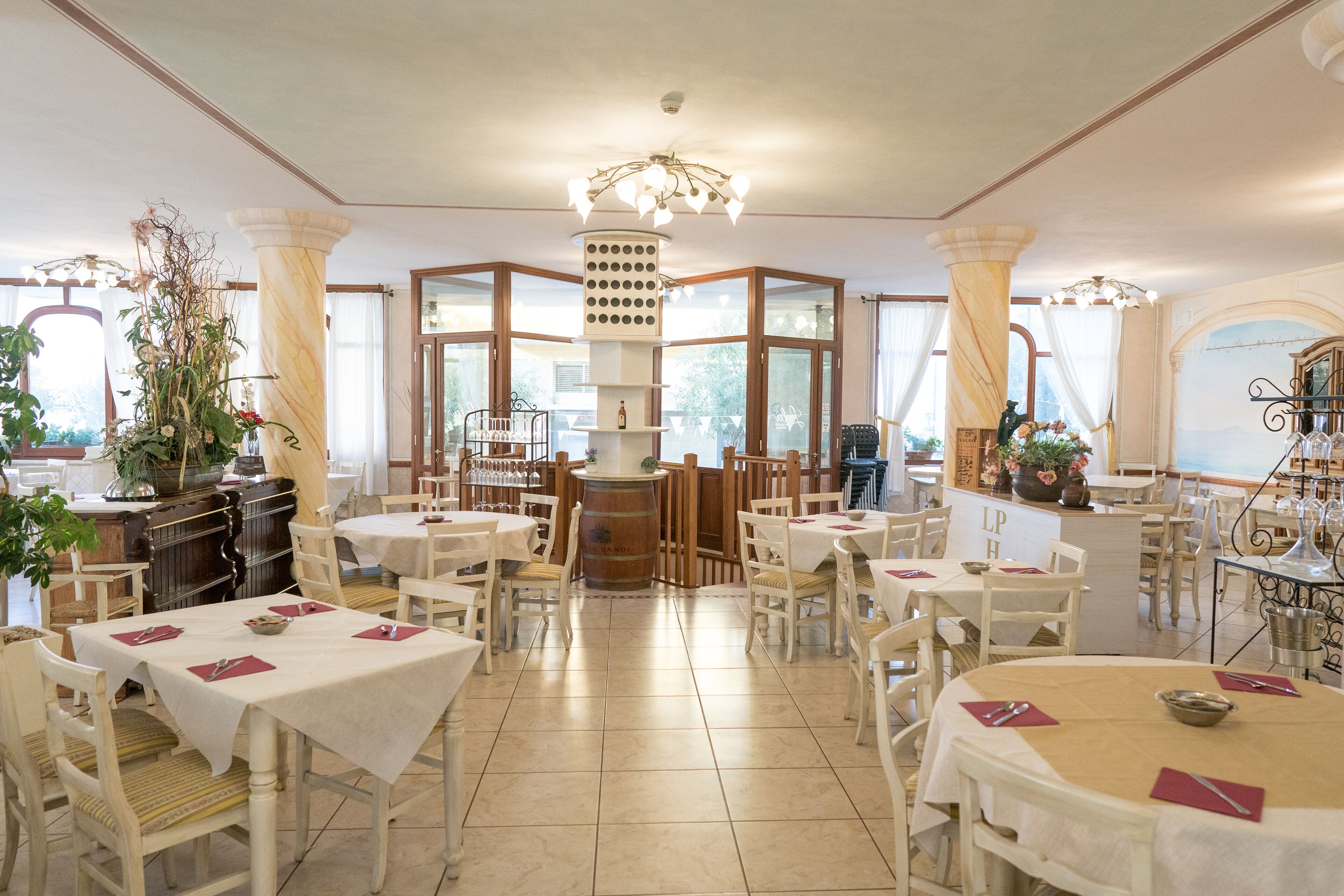 Un'ampia sala da pranzo con diversi tavoli e sedie, una colonna decorativa al centro e grandi finestre che lasciano entrare la luce naturale.