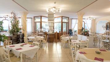 Un'ampia sala da pranzo con diversi tavoli e sedie, una colonna decorativa al centro e grandi finestre che lasciano entrare la luce naturale.