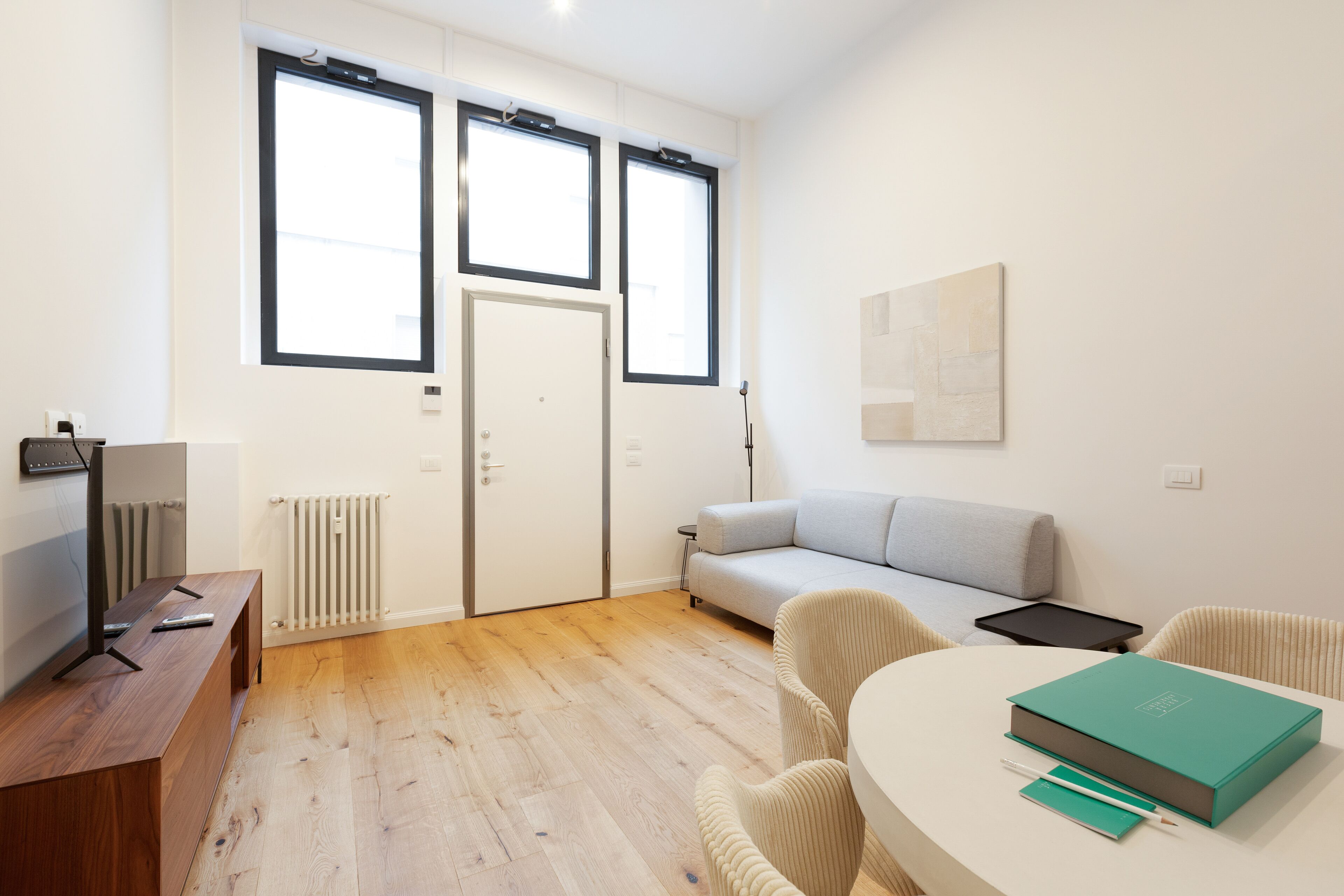 A modern living room with a sofa, a coffee table, and a TV on a wooden stand. There are two windows providing natural light and a radiator under the windows.