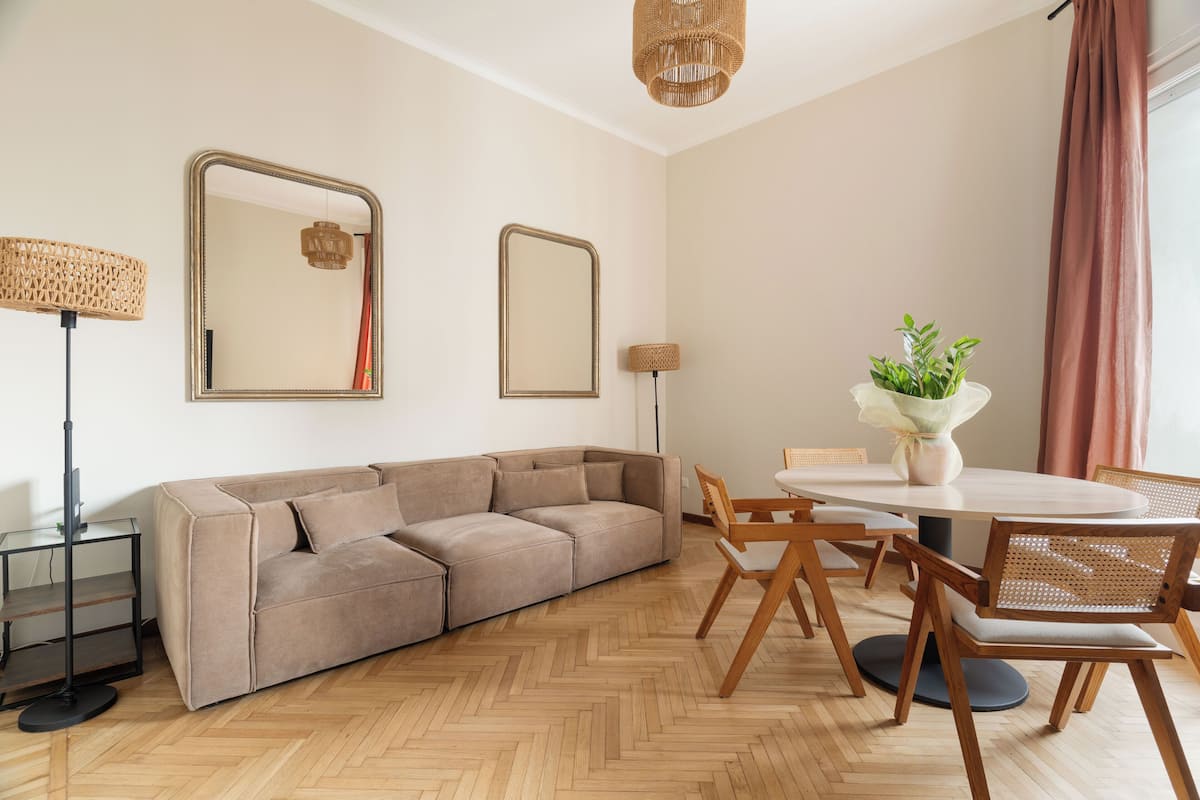 A modern living room with a sectional sofa, round dining table, wooden chairs, and two wall-mounted mirrors.