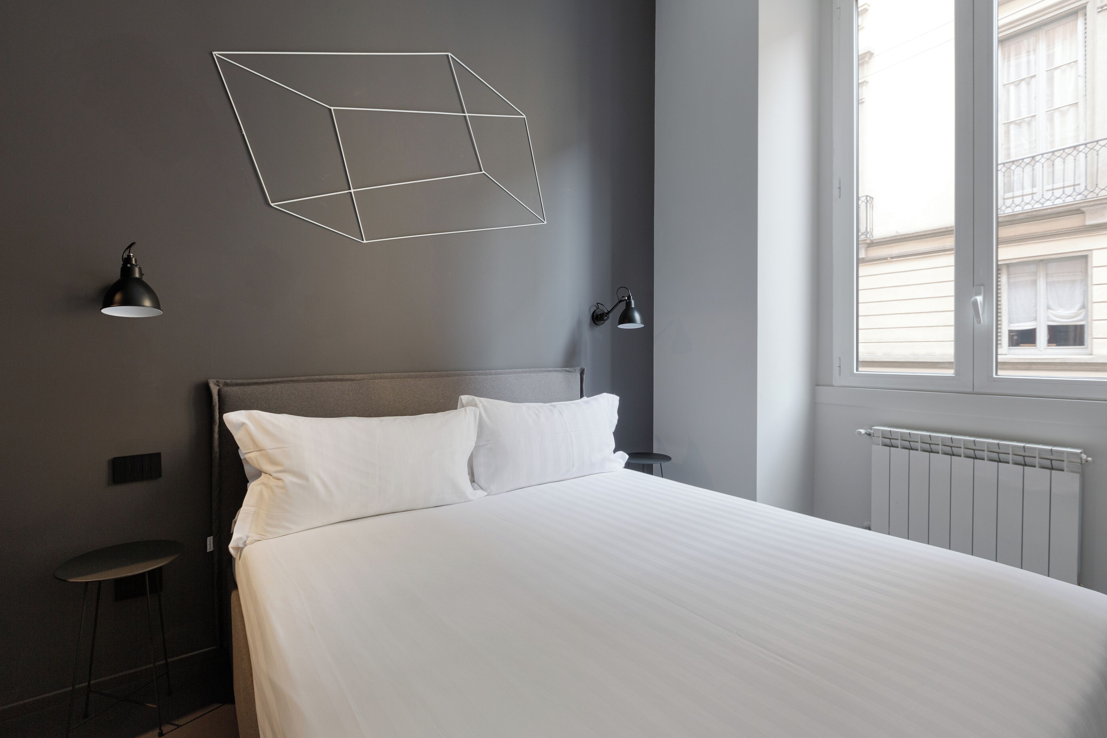 A modern hotel room with a large bed, two bedside lamps, a geometric wall art piece, and a window with a view of buildings.