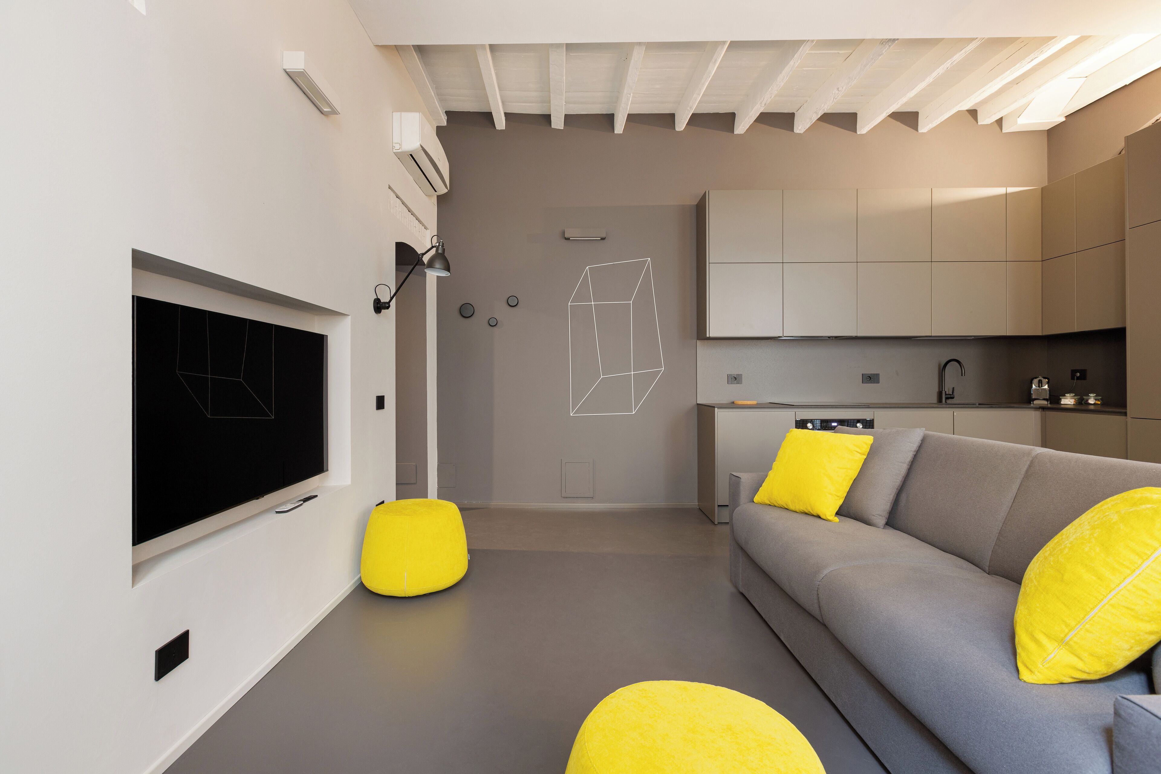 A modern living room with a grey sofa, yellow cushions, a flat-screen TV, and a minimalist kitchen in the background.