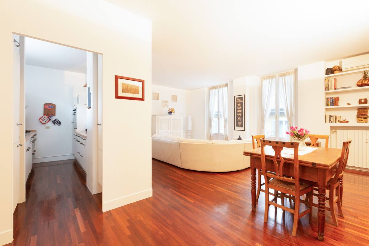A spacious living area with a wooden dining table and chairs, a white sofa, and a bookshelf.