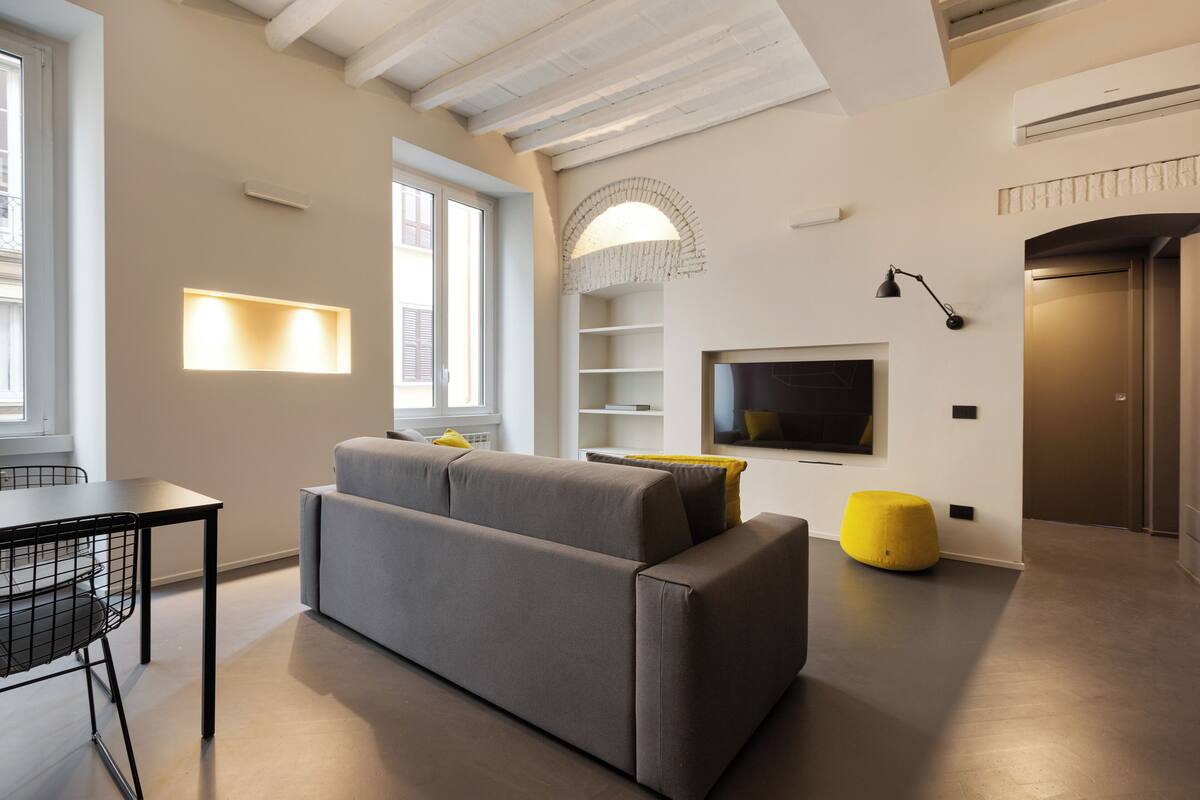 A modern living room with a grey sofa, a yellow ottoman, a black table with chairs, and a TV mounted on the wall.