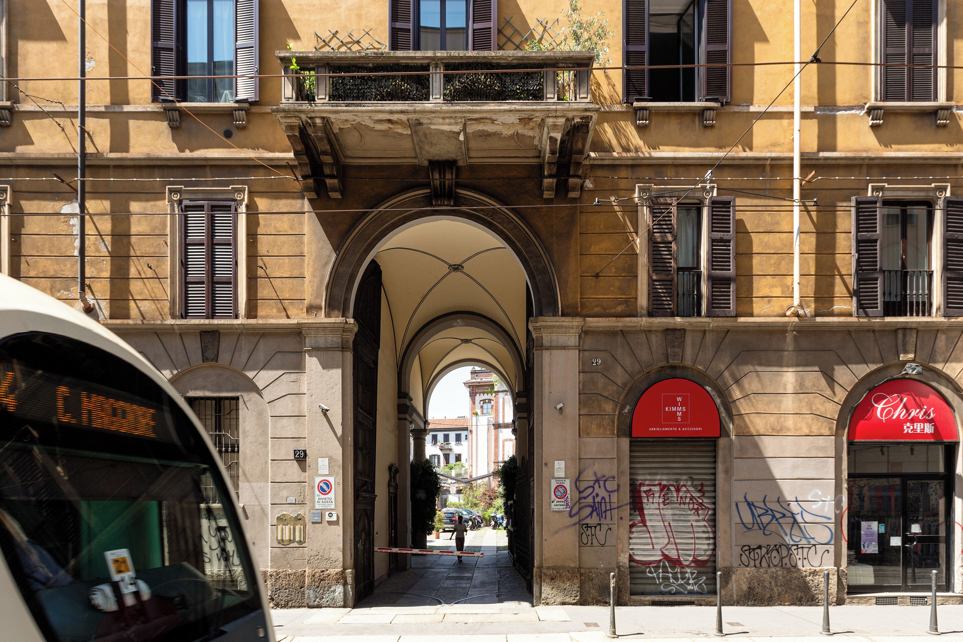 Une vue de rue avec un bâtiment doté d’une entrée en arc de cercle, d’un volet fermé sur la droite et d’un autobus au premier plan.