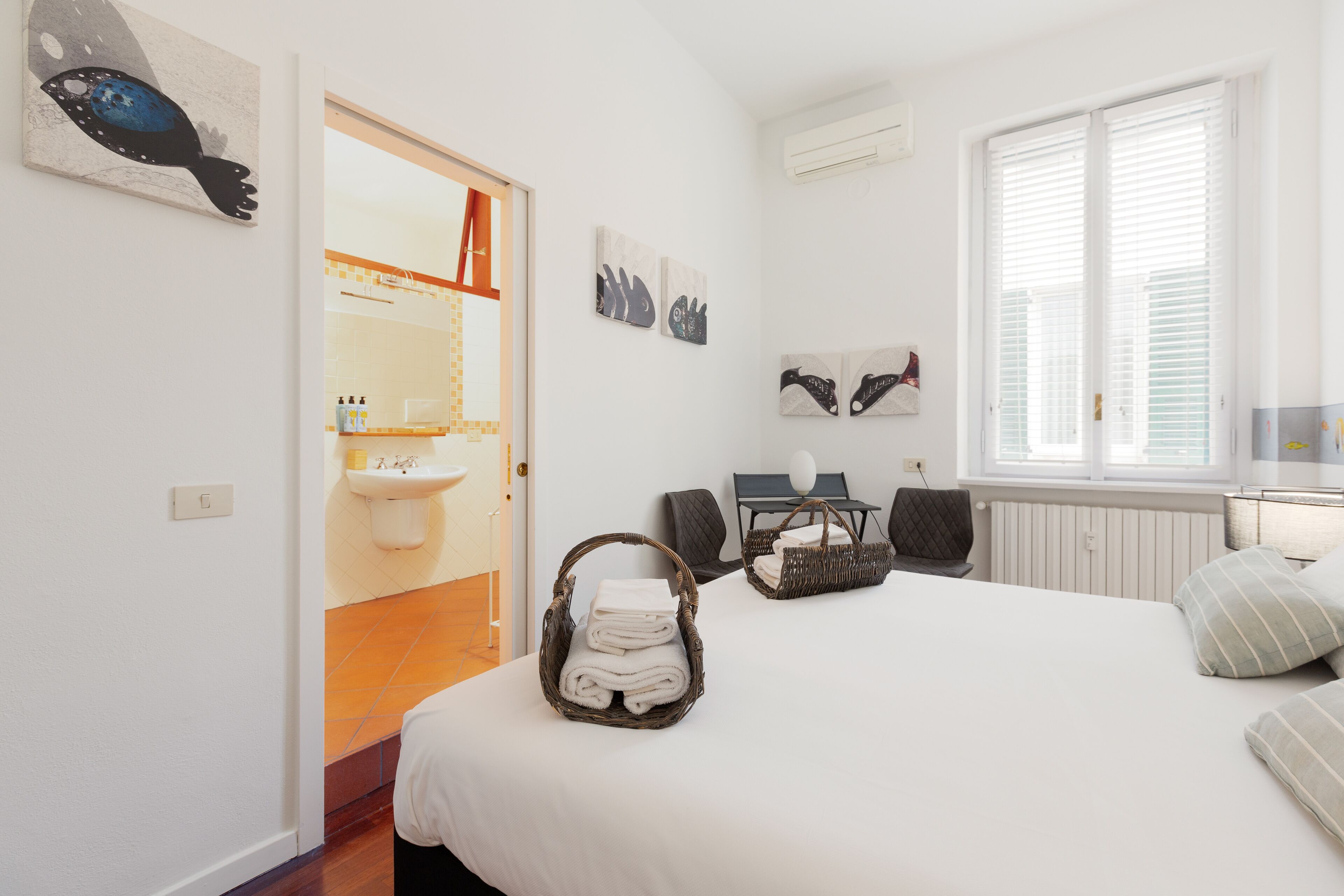 A hotel room with a bed, a desk, a chair, a window with blinds, and artwork on the walls.