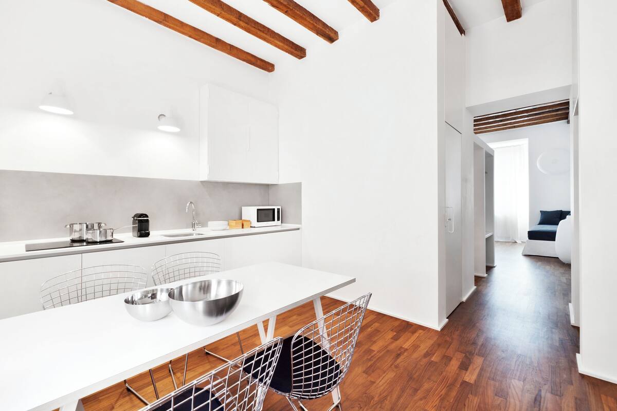 A modern kitchen with white cabinetry, a wooden floor, and a dining area with metal chairs.