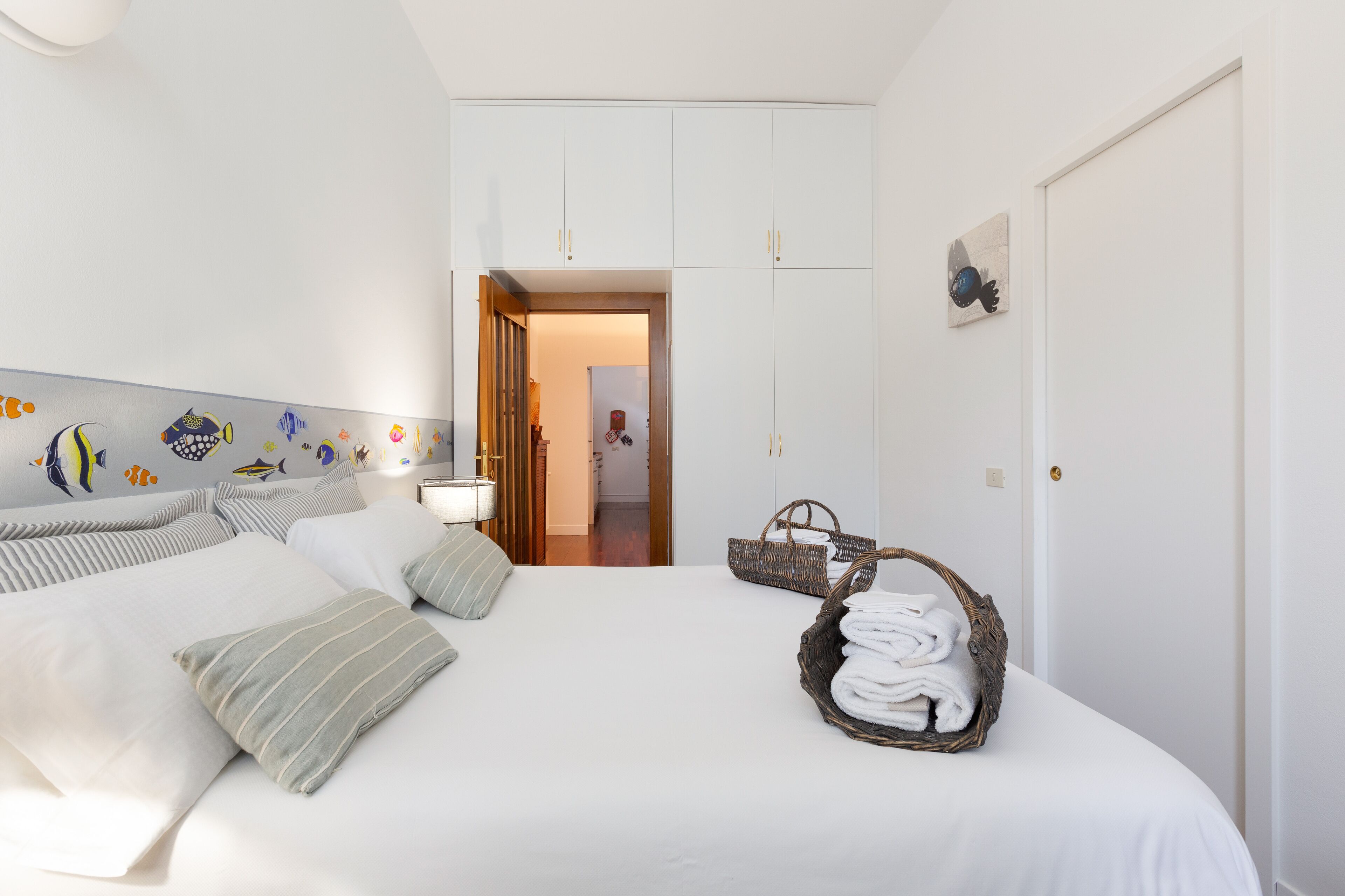Una habitación de hotel con una cama, almohadas, un mural en la pared, una puerta y una cesta de toallas.