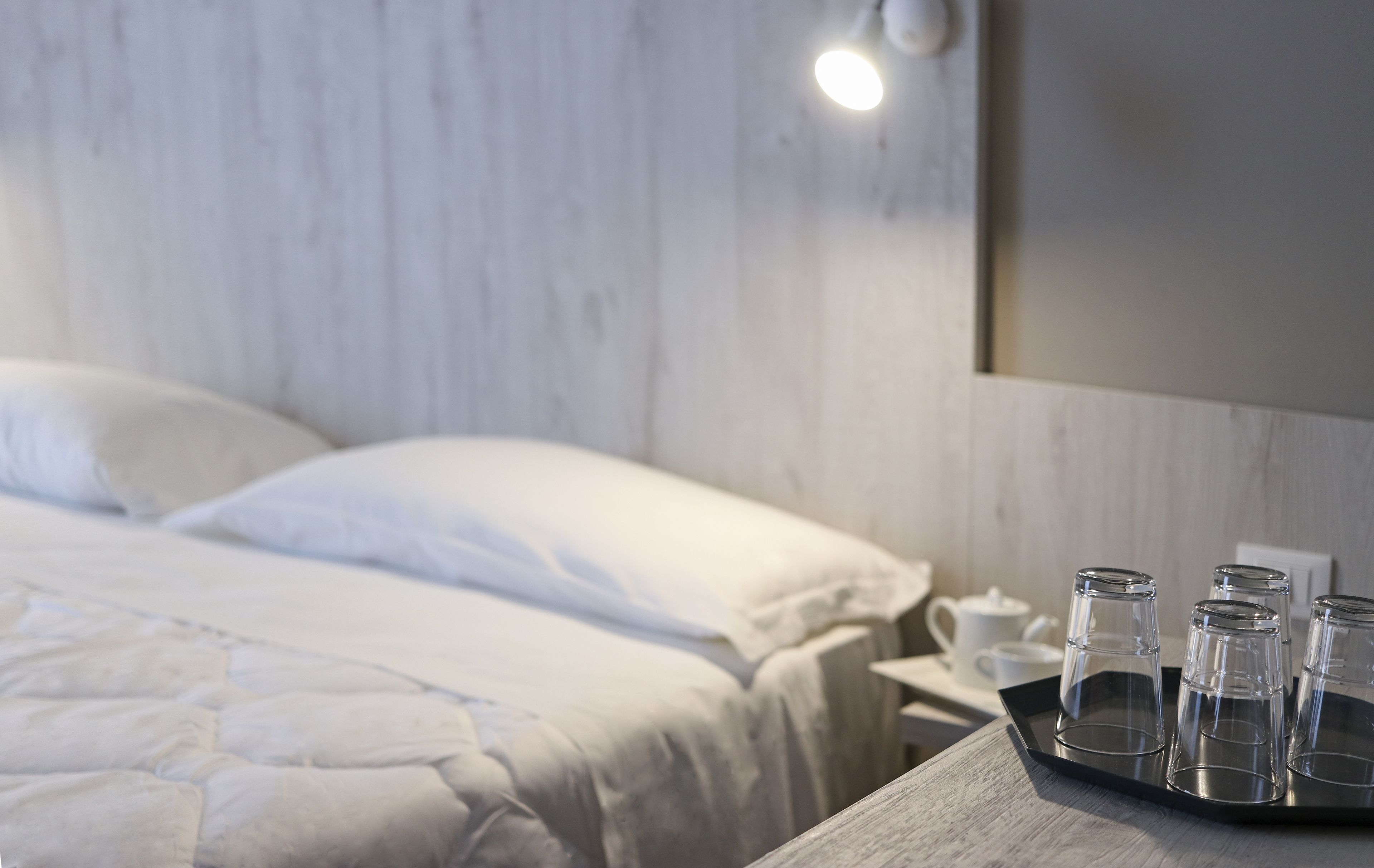 A neatly made bed with white linens, a bedside table with a tray holding four clear glasses, and a white wall with a light fixture.