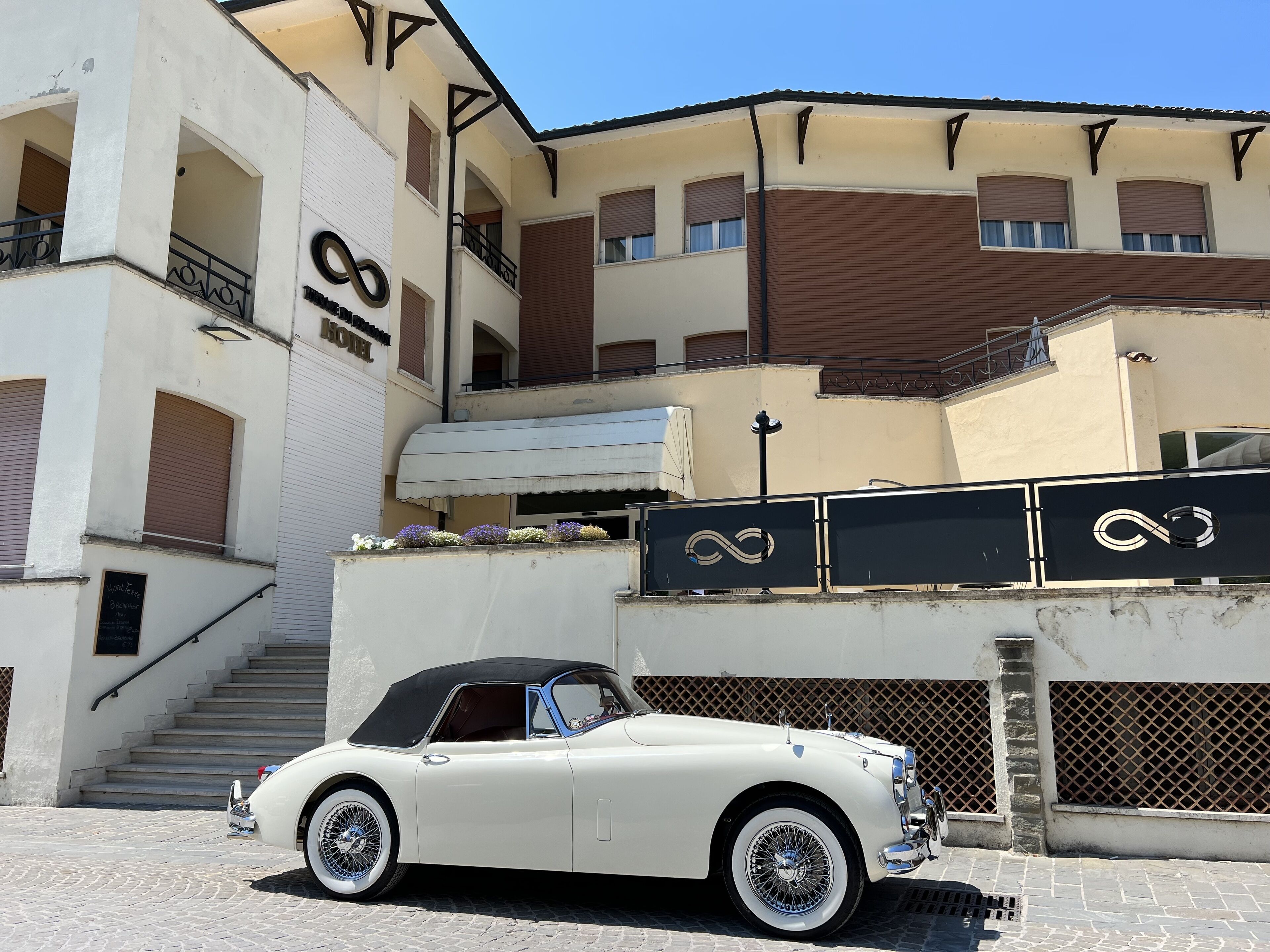 Un coche descapotable blanco antiguo estacionado frente a un hotel con una escalera que lleva a la entrada.
