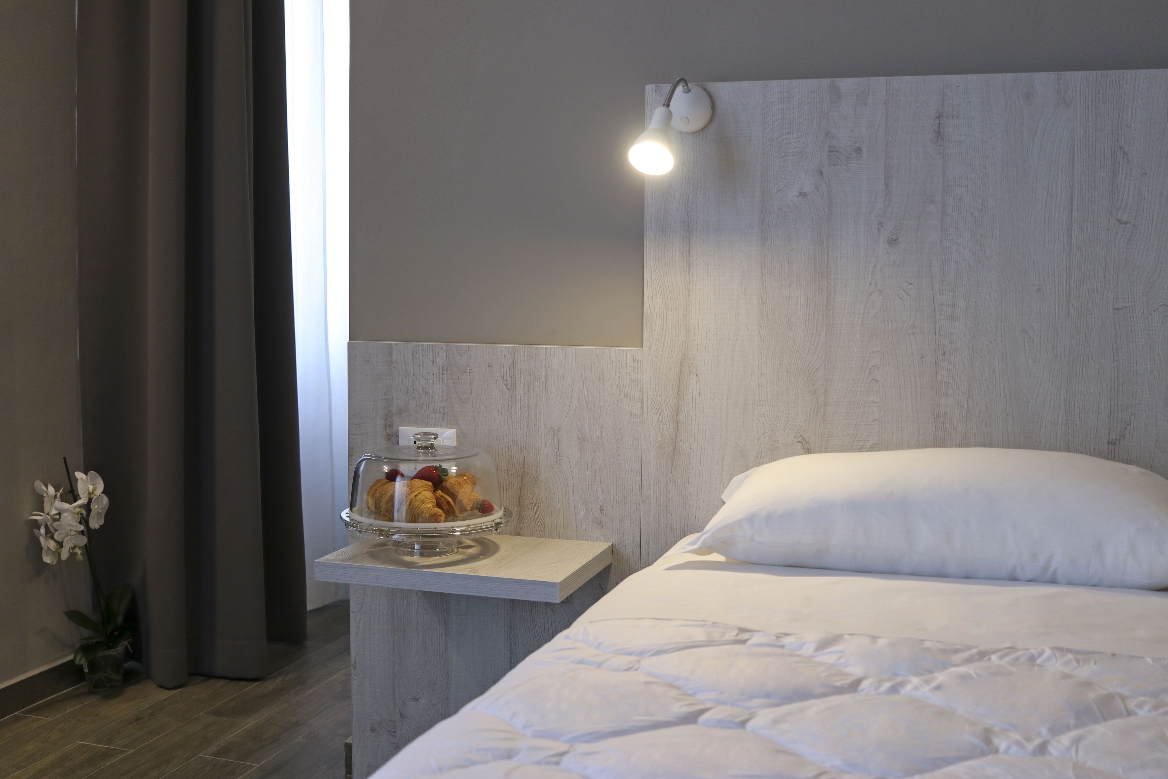 Een hotelkamer met een houten hoofdbord, een nachtkastje met een glazen koepel vol gebak, een wit bed met een sprei en een wandlamp.