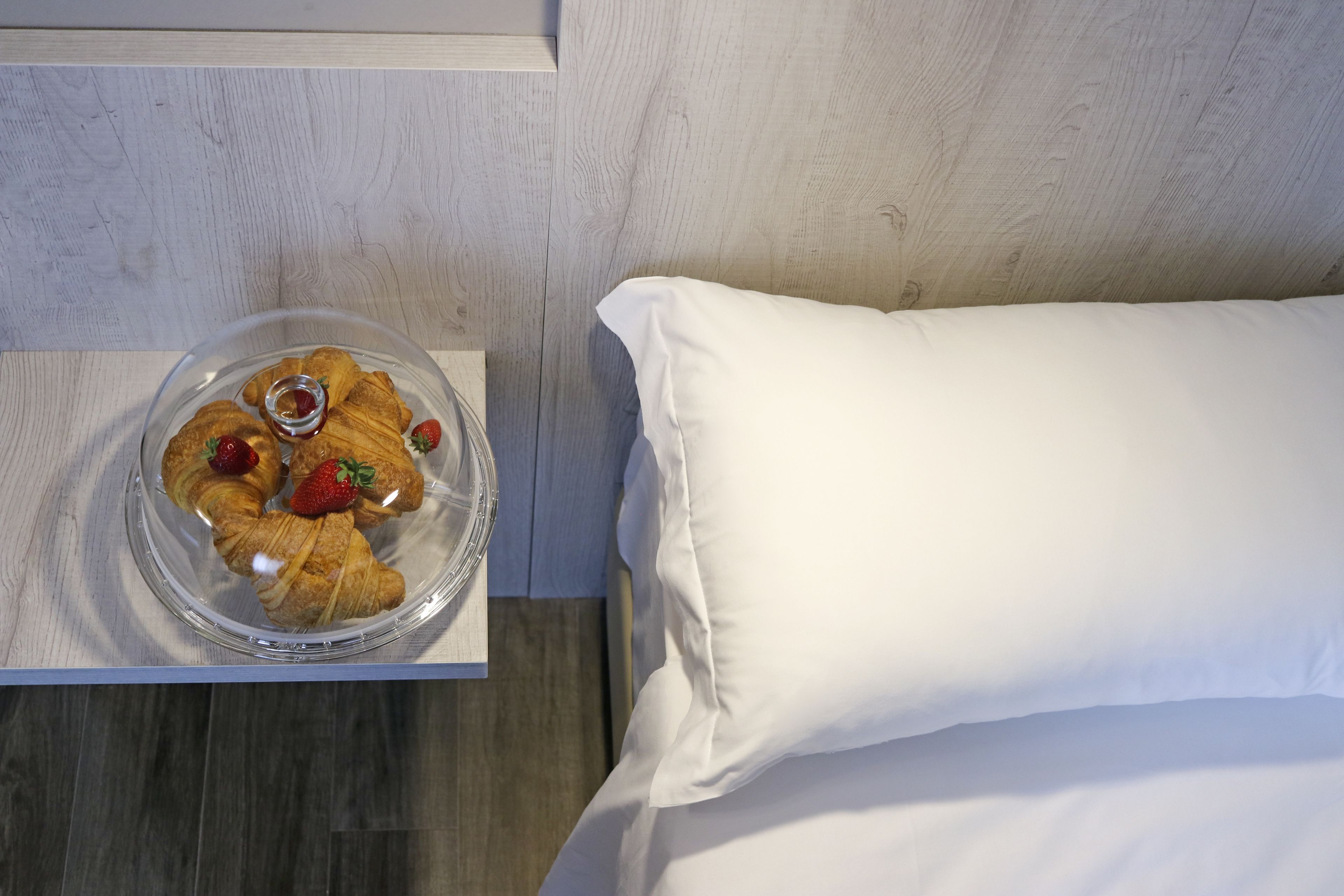 A bed with a white pillow and a clear glass bowl containing croissants and strawberries on a wooden nightstand.