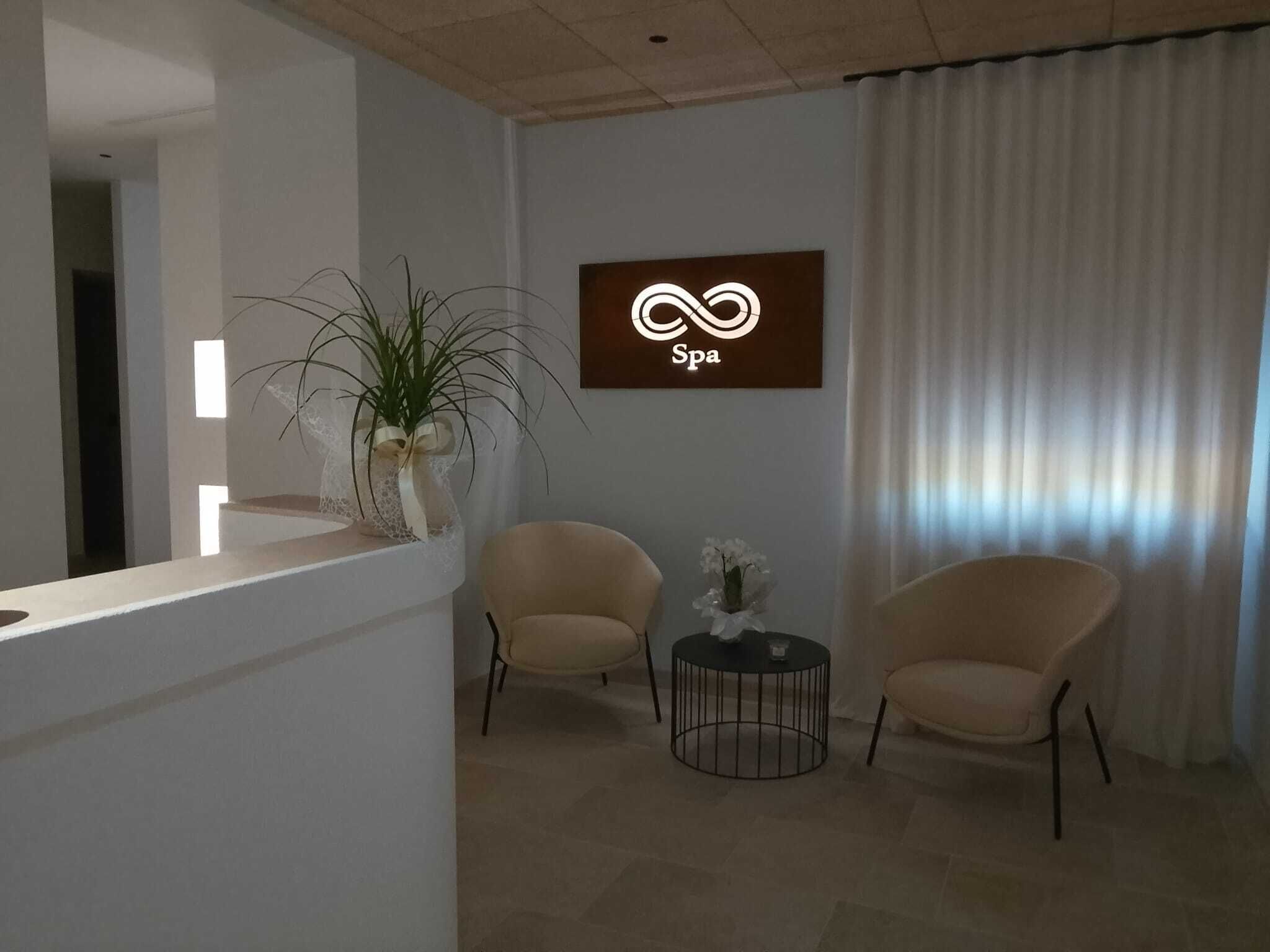 Zona de recepción con mostrador, dos sillones, una mesita con un florero y el logotipo de un spa en la pared.