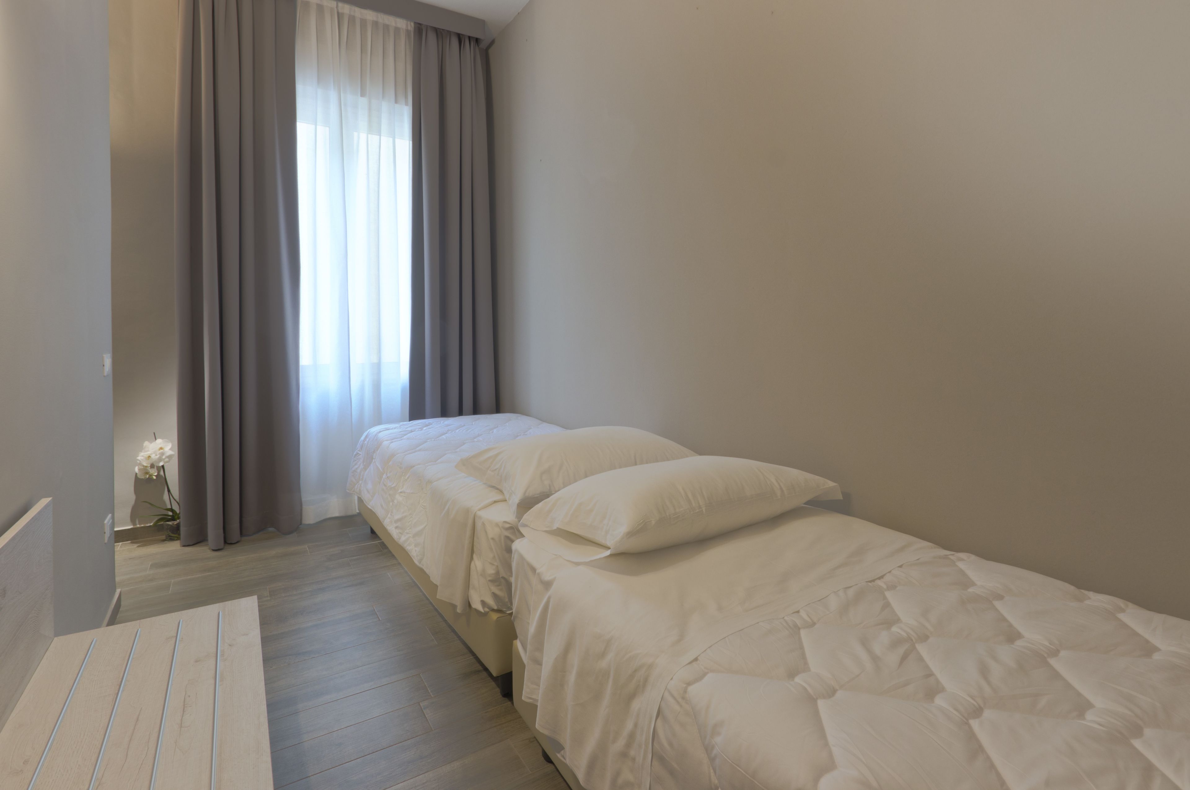 Quarto com uma cama de solteiro, mesa de cabeceira de madeira, um vaso com flores e uma janela com cortinas.