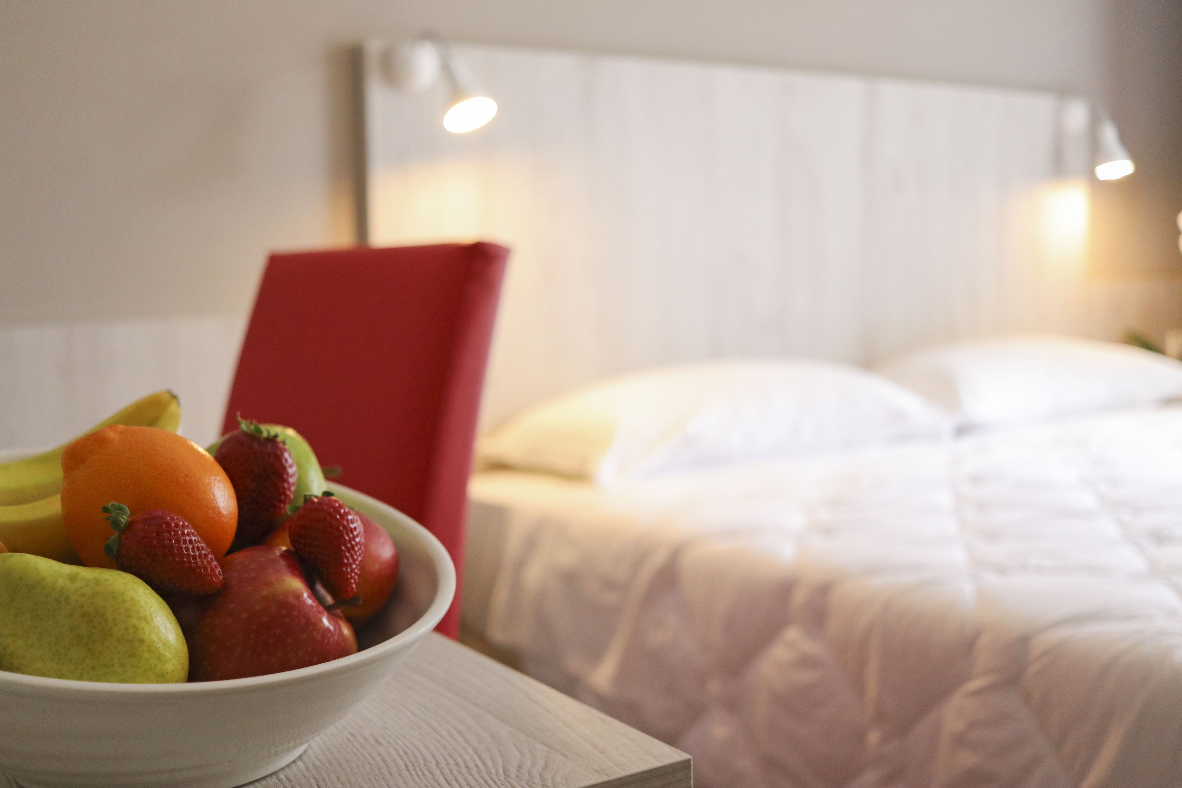 Una cama con ropa de cama blanca, una silla roja y un frutero sobre una mesa.