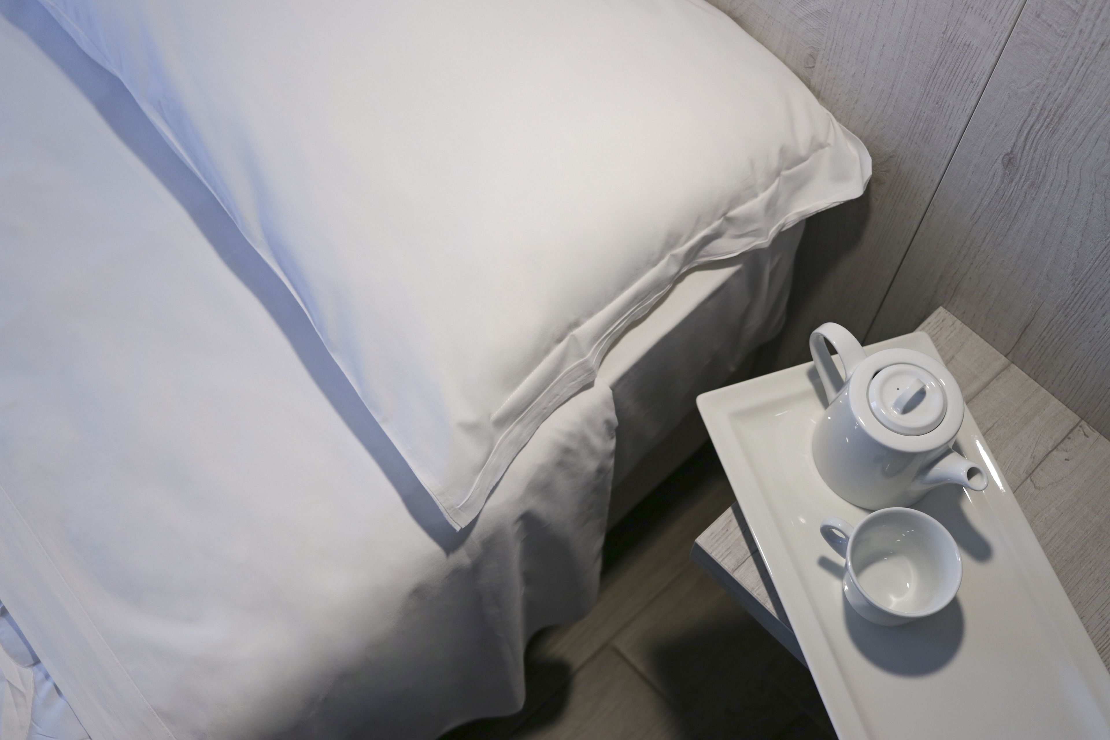 A neatly made bed with white bedding, a white tray with a teapot and cup, and a wooden floor.