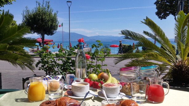 Une table dressée avec des aliments pour le petit-déjeuner, notamment des croissants, des biscuits et des fruits, donnant sur une plage où l’on aperçoit des parasols et des personnes au loin.