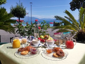 Mesa dispuesta con desayuno, incluyendo croissants, galletas y fruta, con vista a una playa con sombrillas y gente al fondo.