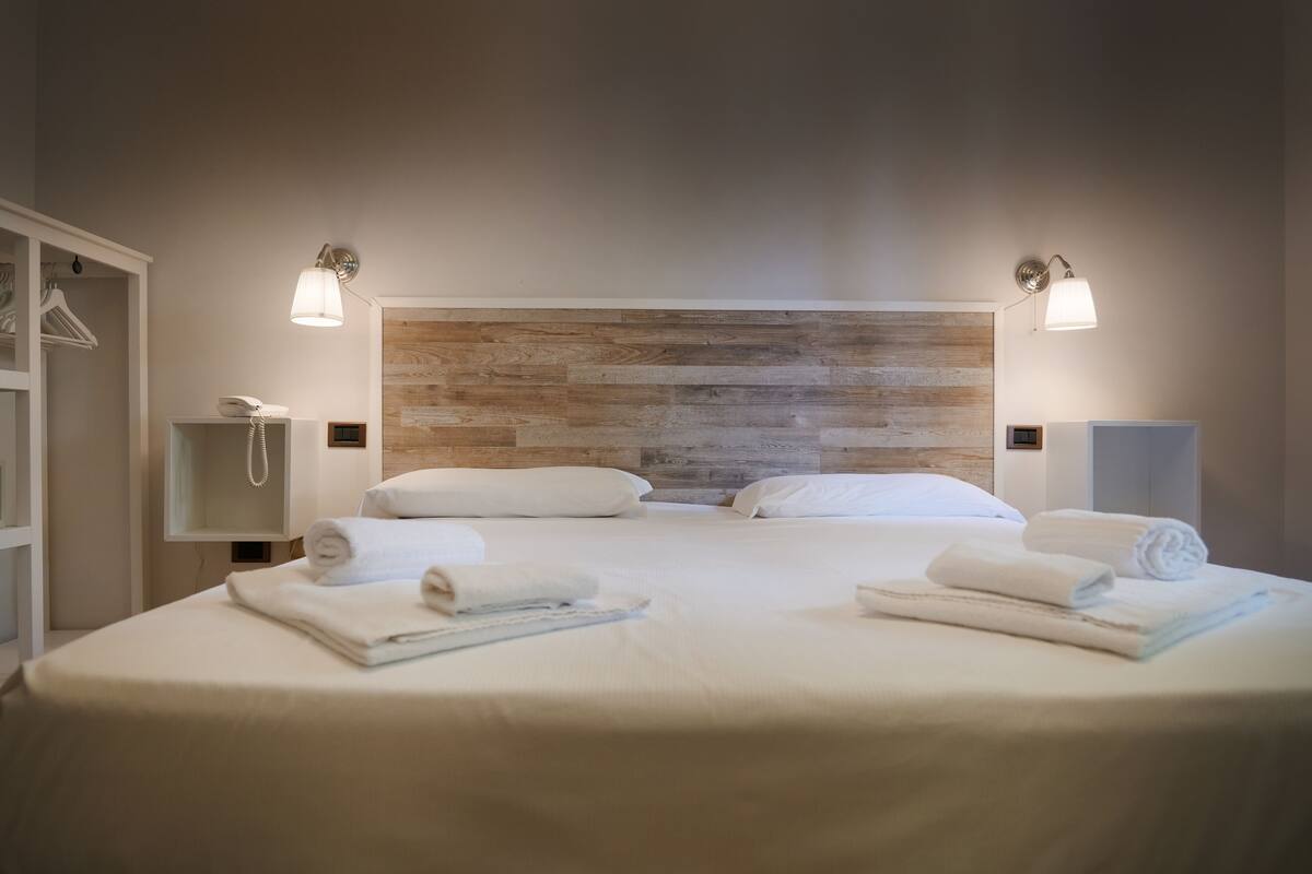 Un lit double avec du linge de lit blanc, deux tables de chevet avec des lampes, une tête de lit en bois et une armoire blanche.