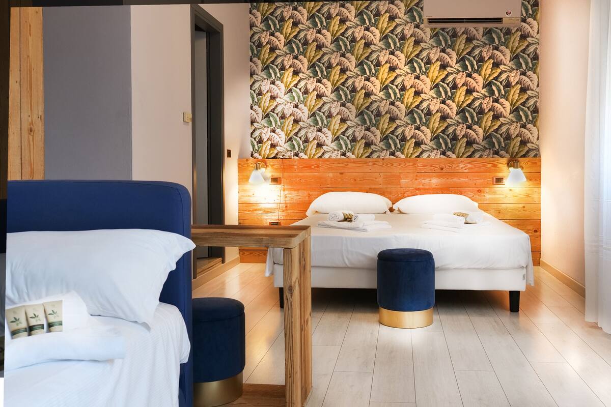 Une chambre d’hôtel moderne avec une tête de lit en bois, un lit avec du linge de maison blanc, un pouf bleu, une table de chevet en bois et un climatiseur fixé au mur.