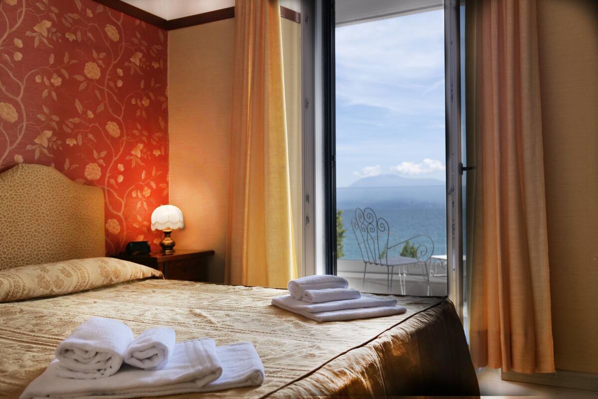 Une chambre d’hôtel avec un lit, une table de chevet, une lampe et une vue sur la mer, visible à travers une grande fenêtre.