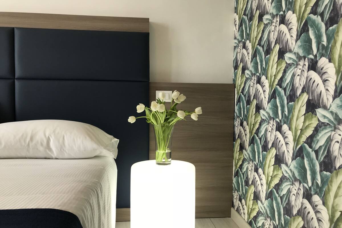 Une chambre moderne avec un lit, une table de chevet ornée d’un vase de fleurs et un mur décoré d’un motif de feuilles tropicales.