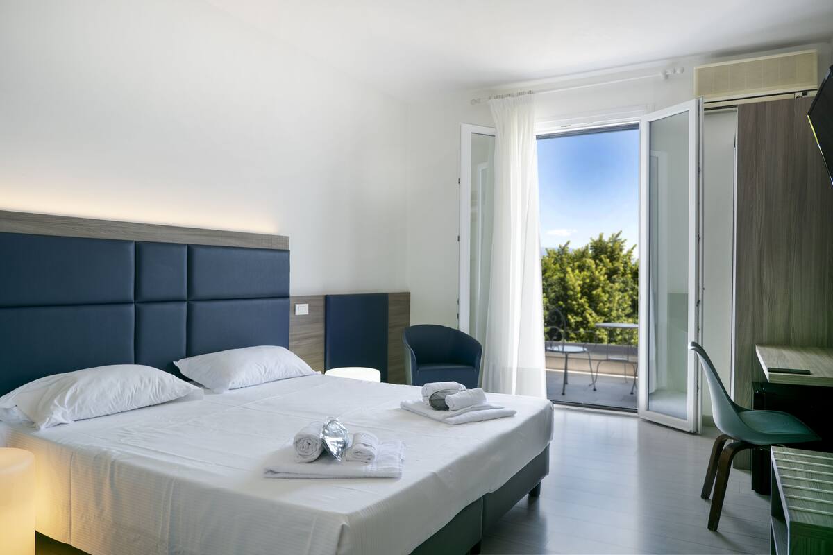 Une chambre d’hôtel moderne avec un grand lit, un bureau et un balcon donnant sur la verdure.