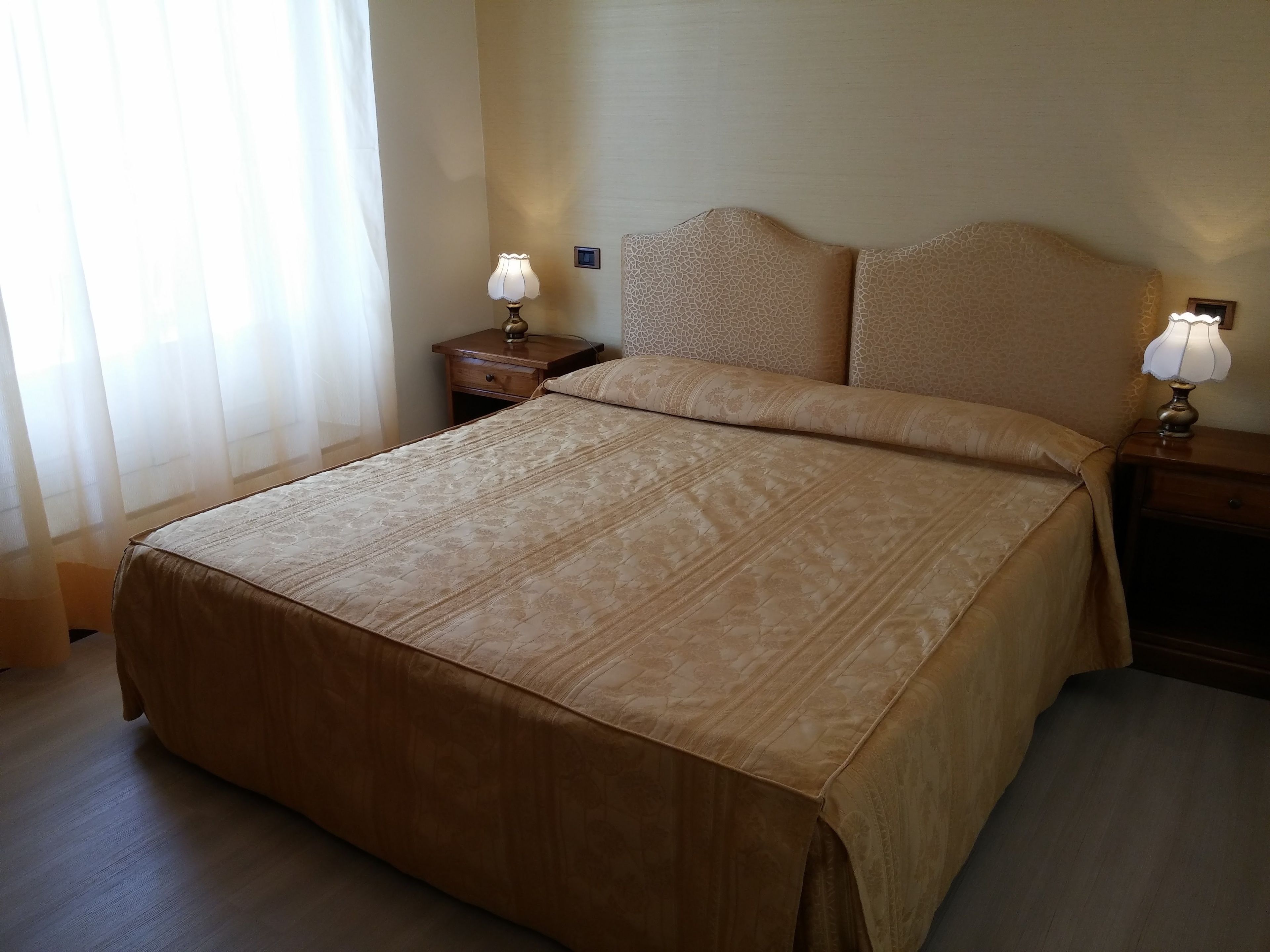 A hotel room with a large bed, two bedside tables with lamps, and a window with sheer curtains.