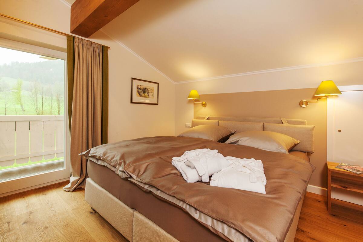 A bedroom with a large bed, two bedside lamps, a window with curtains, and a framed picture on the wall.