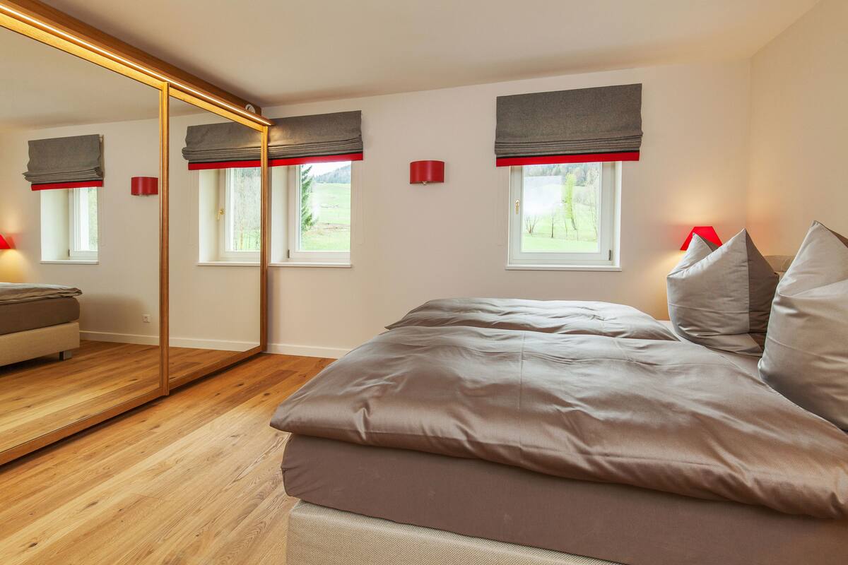 A bedroom with a large bed, a mirror, two windows with red curtains, and a wooden floor.