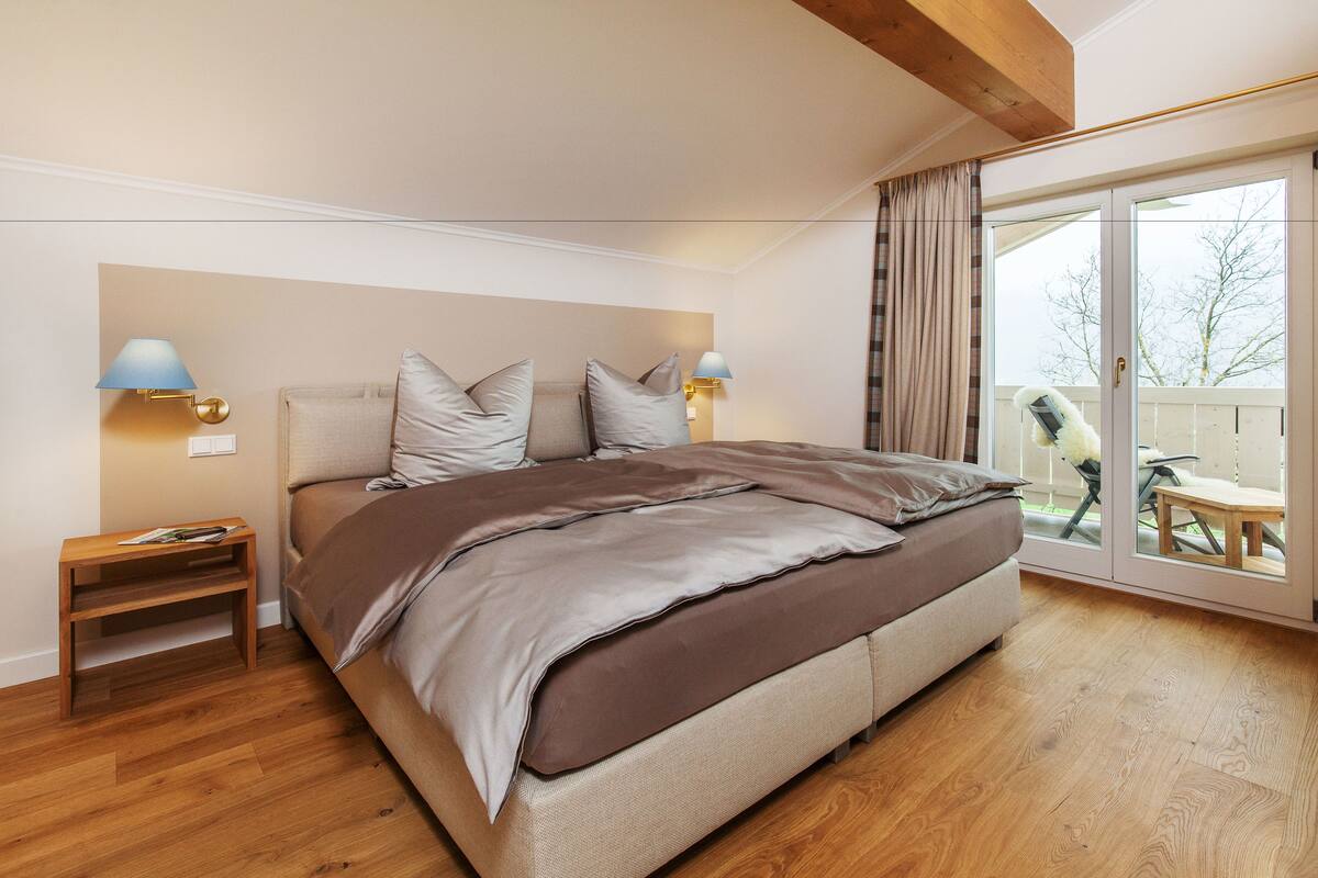 A bedroom with a large bed, wooden floor, two bedside tables with lamps, and a sliding glass door leading to an outdoor area.