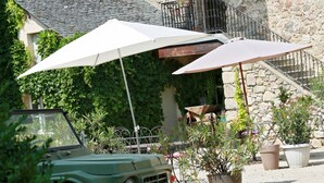Une voiture ancienne de couleur verte est garée devant un bâtiment en pierre doté d’un balcon et d’un espace extérieur aménagé pour s’asseoir.