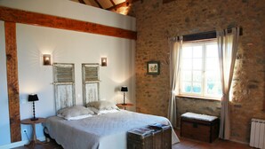 Une chambre avec un grand lit, deux lampes de chevet, un mur en pierre, une fenêtre avec des rideaux et un petit banc en bois.