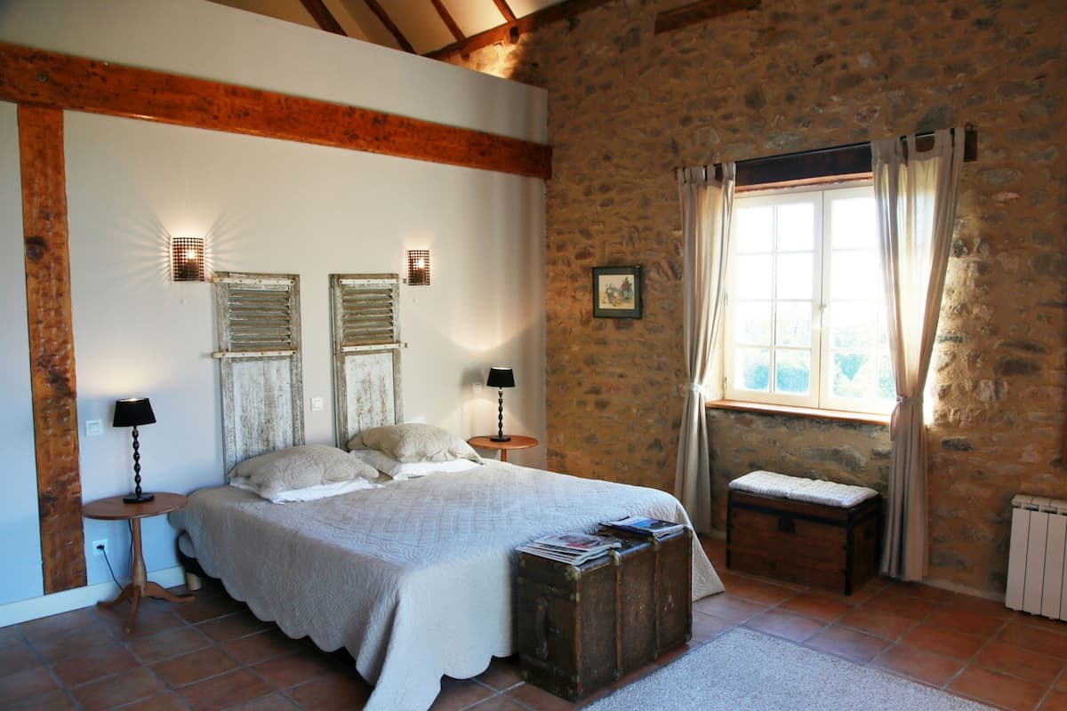 A bedroom with a large bed, two bedside lamps, a stone wall, a window with curtains, and a small wooden bench.