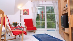 A room with a dining table, chairs, a television, and a large window with curtains. - Weingut Sauer-Kettermann (Enkirch)