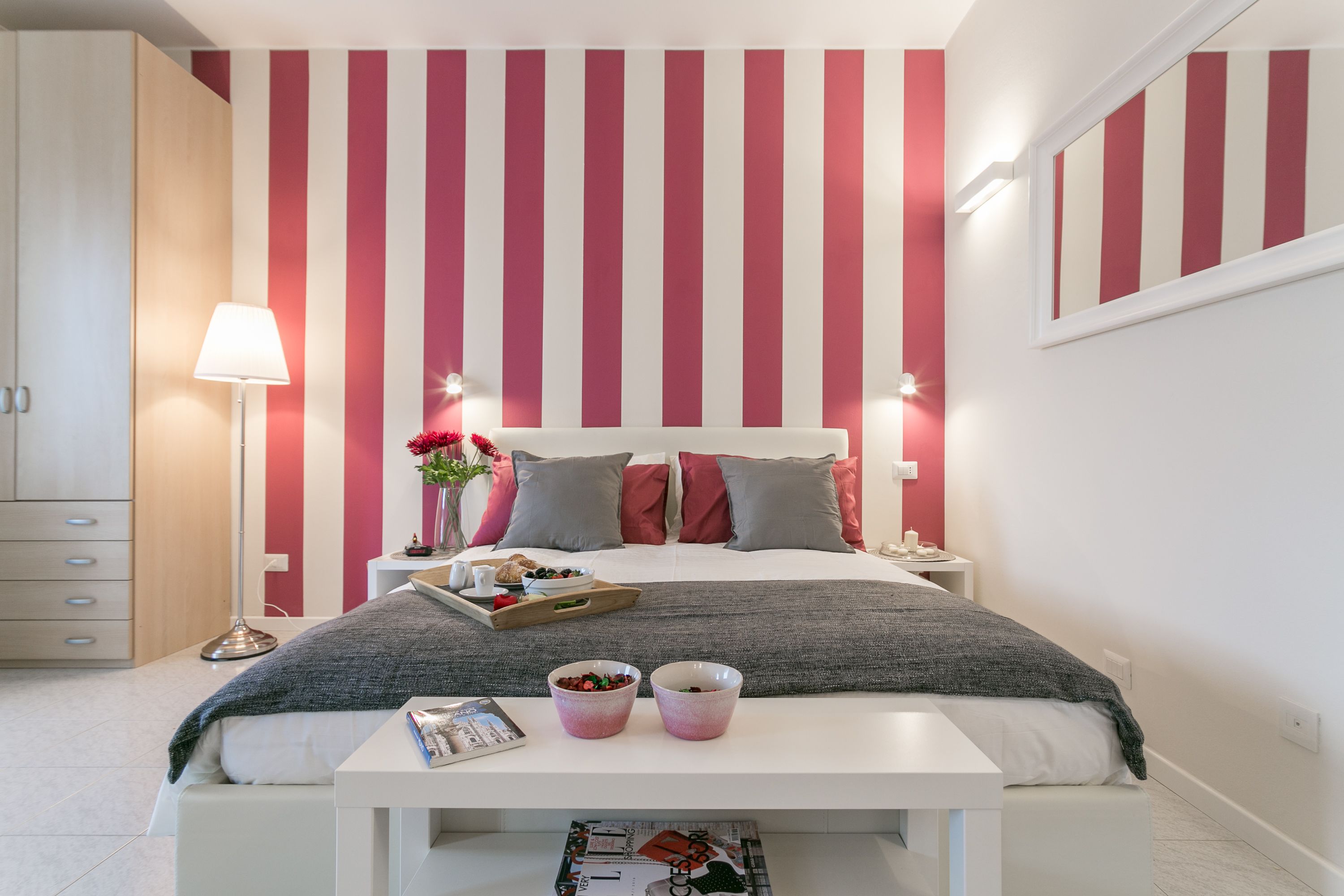 A bedroom with a striped wall, a bed with pillows, a bedside table with a lamp, and a tray with food.