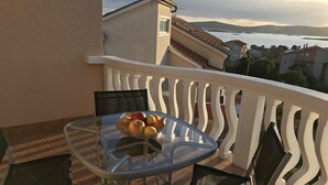 A balcony with a glass table holding fruit, black chairs, and a view of a body of water and buildings in the distance.