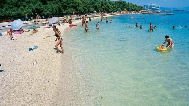 A beach with clear water, people swimming and sunbathing, and a forested area in the background.