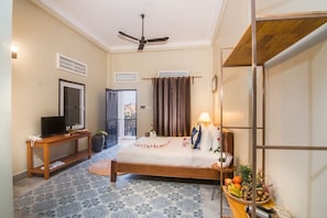 Habitación de hotel con cama, televisión, ventilador de techo, una mesita con frutas y vistas al exterior.
