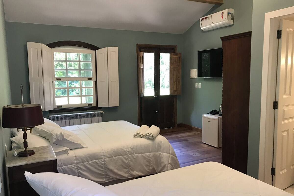 A bedroom with two beds, a nightstand with a lamp, a window with shutters, a door, and a small cabinet.
