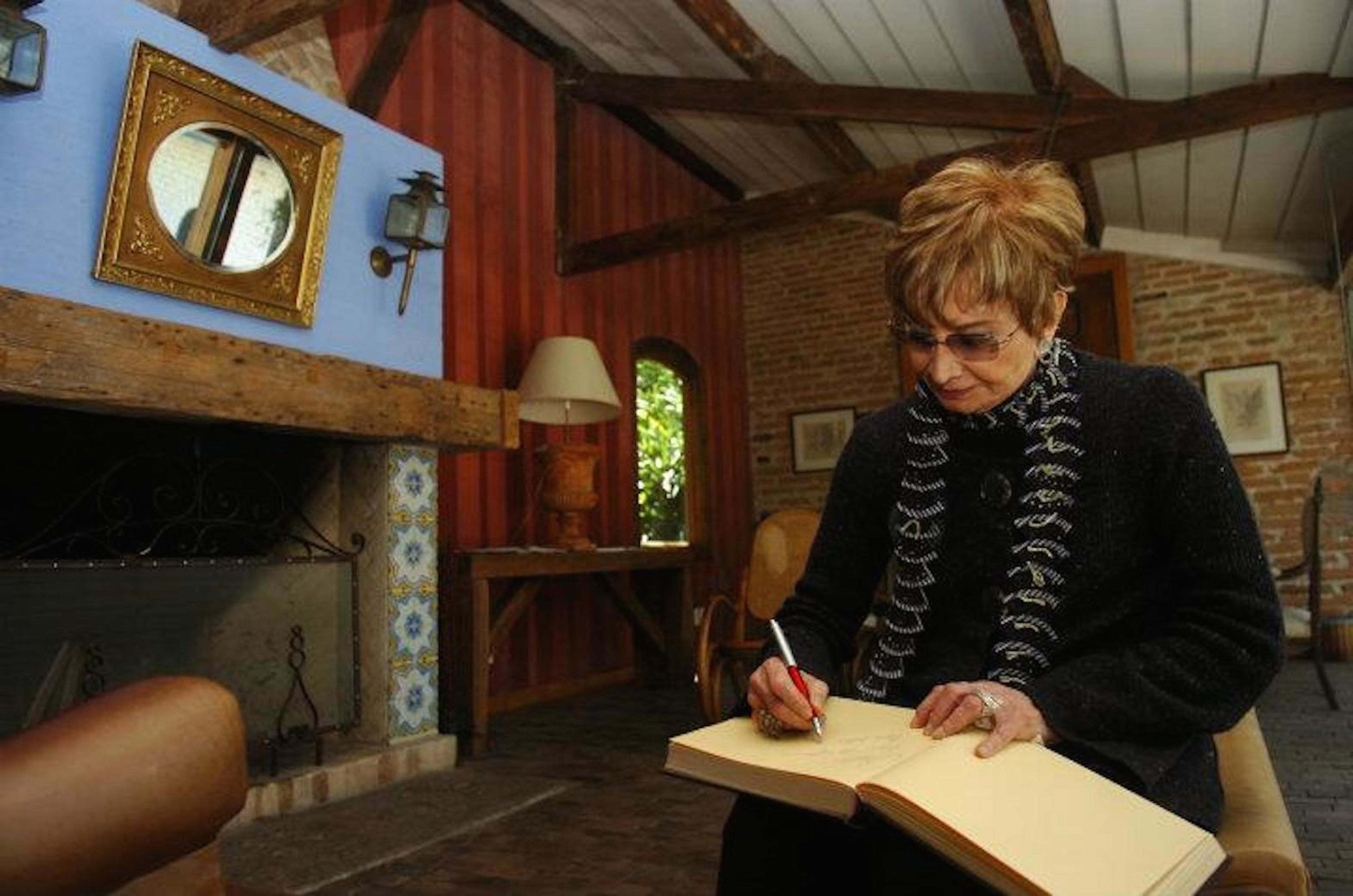 Una persona escribiendo en un libro en una habitación con chimenea, vigas de madera y un espejo en la pared.