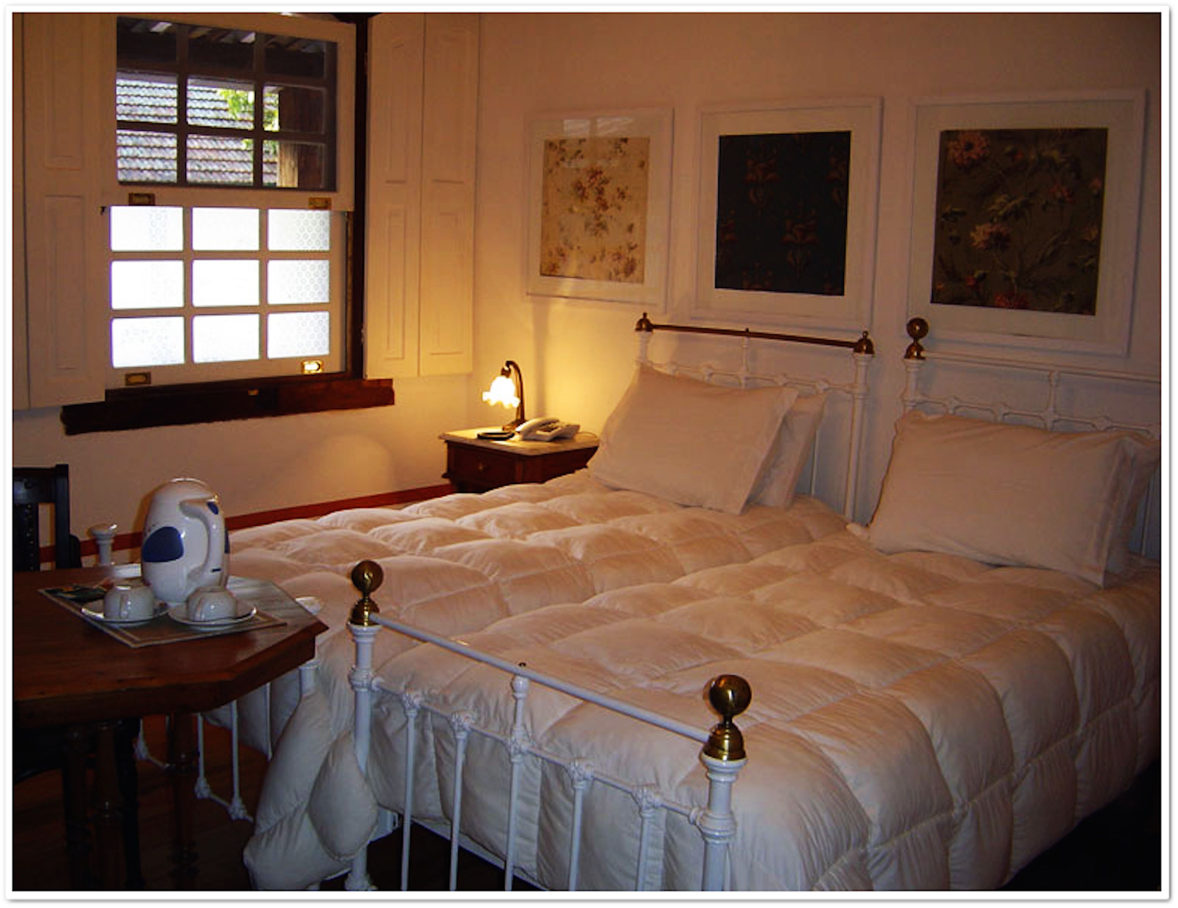 A neatly made bed with a white comforter and pillows, a bedside table with a lamp and a telephone, and a window with a view of greenery.