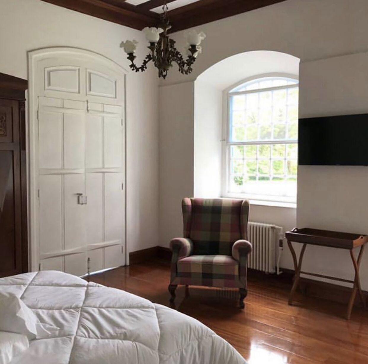 Una habitación con cama, un sillón a cuadros, una puerta blanca, una ventana con vistas a la vegetación y un radiador.