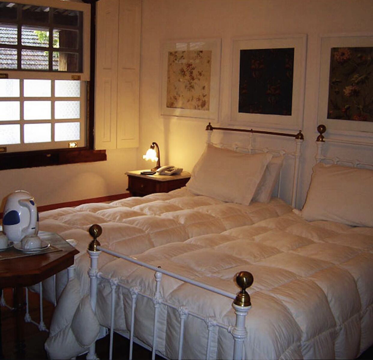 Una cama bien hecha con cabecera y piecera blancos, una mesita de noche con lámpara, y una ventana con persianas.