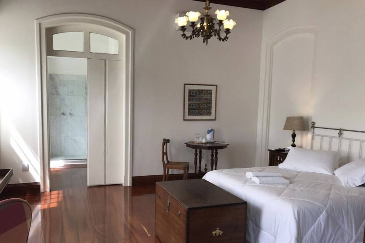 A bedroom with a bed, a wooden chest, a desk with a chair, a chandelier, and a door leading to another room.