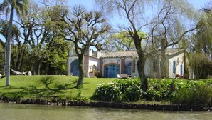 A riverside property with a building featuring a tiled roof and arched windows, surrounded by lush greenery and trees. - Charqueada Santa Rita Pousada de Charme (Pelotas)