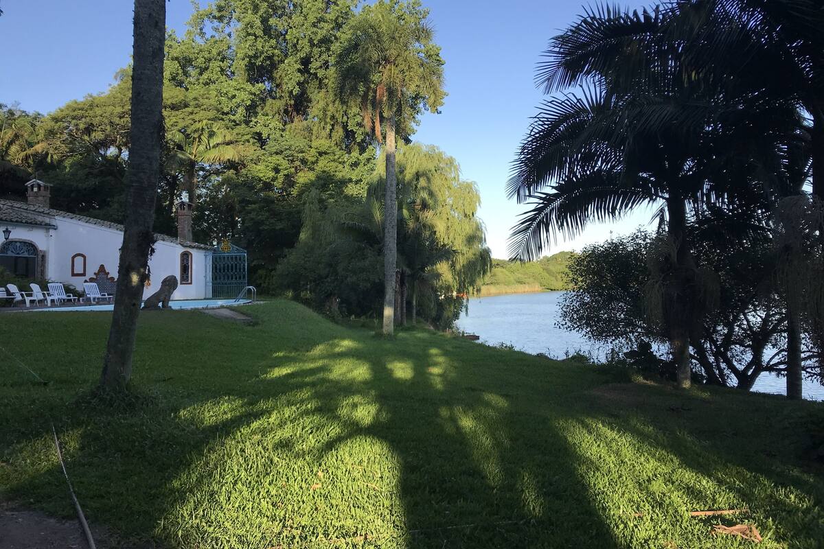 A serene riverside setting with lush greenery, a white building with a pool, and a clear blue sky.