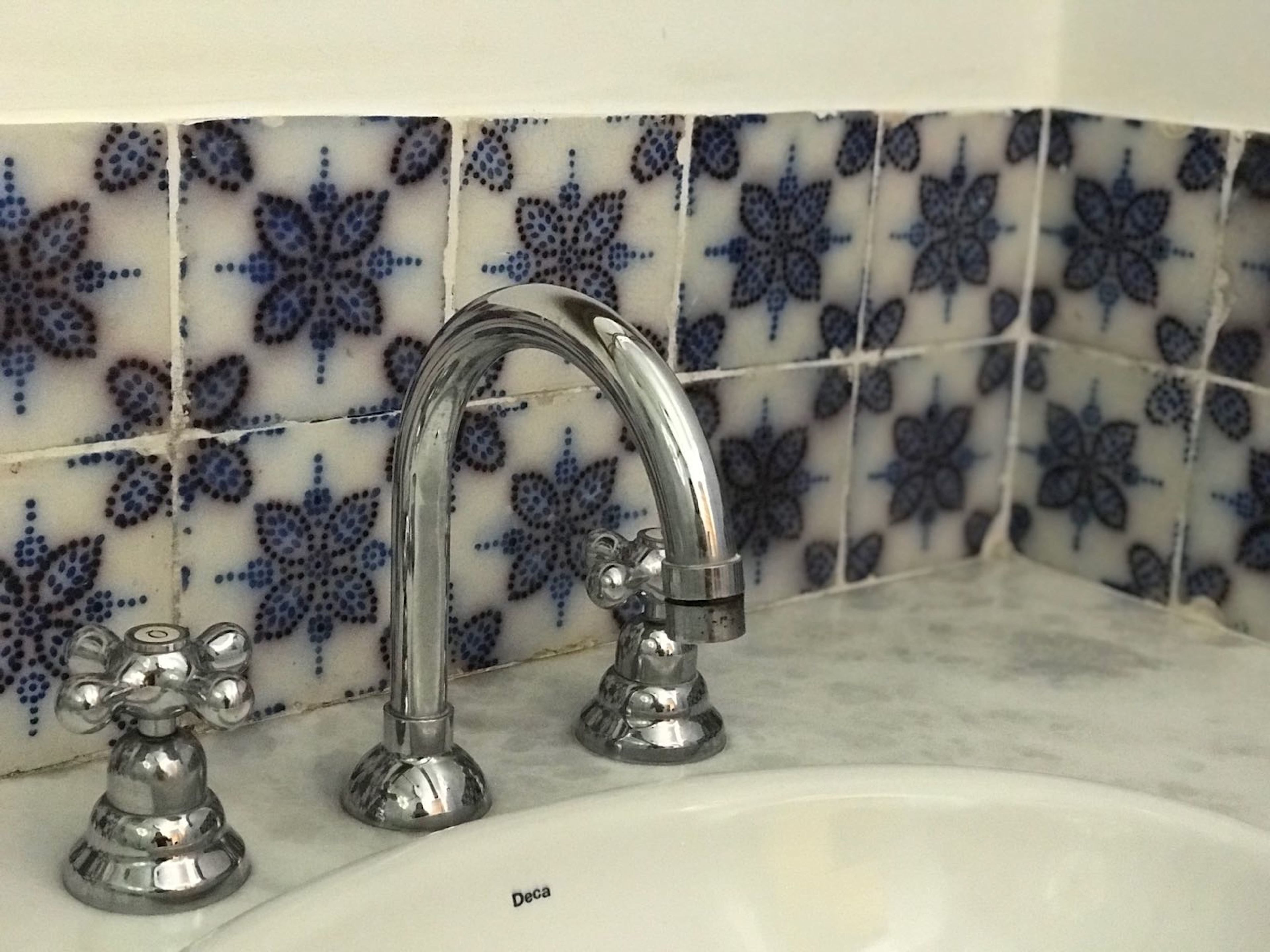 Un baño con un lavamanos blanco y dos grifos cromados. La pared detrás del lavamanos tiene azulejos blancos y azules con motivos florales.