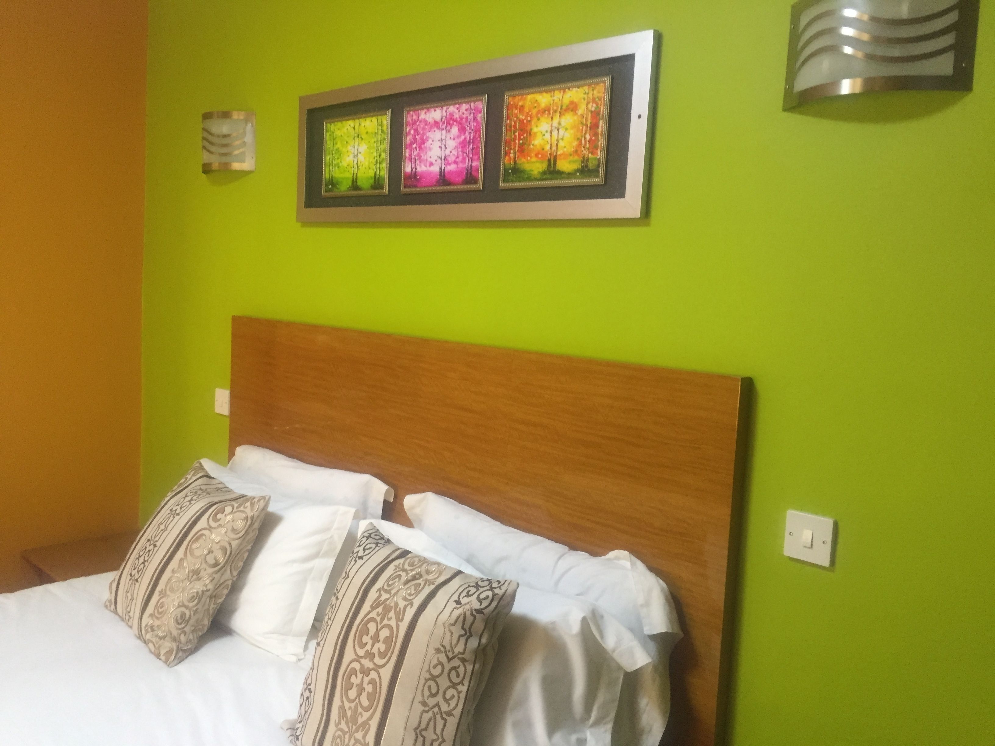 Un lit avec du linge de lit blanc et des oreillers à motifs, une tête de lit en bois et un mur vert orné d’œuvres d’art encadrées.