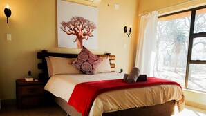 A bedroom with a bed, a nightstand, a wall-mounted lamp, a window with curtains, and a framed picture on the wall. - Zingela Nature Reserve - Leadwood Lodge (Blouberg)